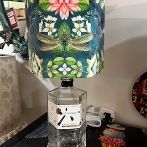 May include: A distinctive table lamp crafted from a recycled gin bottle. The lampshade is cylindrical, with a teal backdrop and a pattern of flowers and dragonflies. The bottle displays a "ROKU GIN" label.