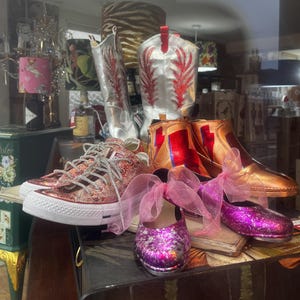 May include: A display of various shoes: silver cowboy boots with red detailing, gold and red ankle boots, pink glitter Converse trainers, and purple glitter baby shoes. The footwear is arranged on a surface.
