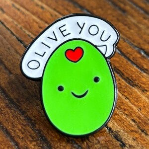 May include: A green enamel pin in the shape of an olive, featuring a smiling face and a red heart. Above the olive is a white banner with the words "Olive You" in black. The pin is set against a wooden background.