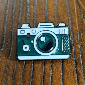 May include: A green and white enamel pin badge in the shape of a vintage camera. The badge features a detailed design with a lens, buttons, and other camera elements. The badge is set against a wooden background.