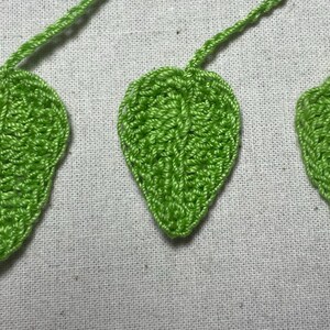 May include: Three handmade crochet leaves in varying sizes. The leaves are a vibrant green color and feature detailed stitching, with stems extending from the top. The leaves are displayed on a light-colored fabric background.