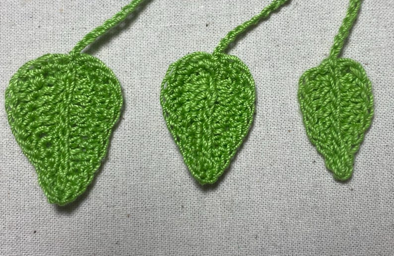 May include: Three handmade crochet leaves in varying sizes. The leaves are a vibrant green color and feature detailed stitching, with stems extending from the top. The leaves are displayed on a light-colored fabric background.