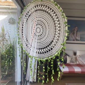 May include: A large, circular white crochet dream catcher with intricate patterns. The dream catcher is adorned with green leaf-shaped embellishments around the edge and hanging down. It hangs in front of a window, with a glimpse of a room in the background.