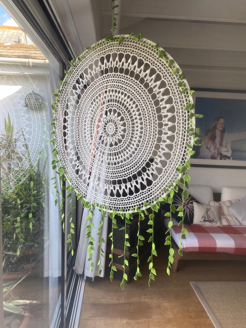 May include: A large, circular white crochet dream catcher with intricate patterns. The dream catcher is adorned with green leaf-shaped embellishments around the edge and hanging down. It hangs in front of a window, with a glimpse of a room in the background.