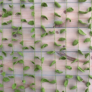 May include: A handmade crochet curtain with green leaves and vines. The leaves are spaced along vertical green strings, creating a natural design. The background is a light-coloured wooden surface.