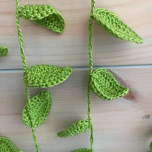 May include: Close-up of a handmade crochet vine with bright green leaves. The leaves are detailed with textured stitching and are attached to a twisted green stem. The background is a light-colored wooden surface.