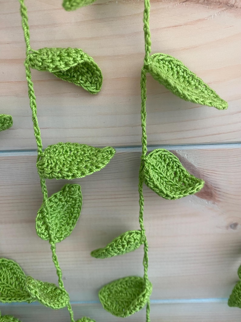 May include: Close-up of a handmade crochet vine with bright green leaves. The leaves are detailed with textured stitching and are attached to a twisted green stem. The background is a light-colored wooden surface.
