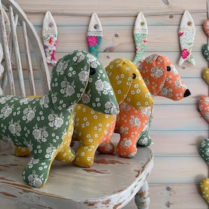 Puede incluir: Tres juguetes de tela con forma de perro salchicha con estampados florales verdes, amarillos y naranjas están sentados en una silla de madera blanca. Adornos decorativos con forma de pez y corazón cuelgan de la pared detrás de ellos. Los perros tienen ojos de botón negros.