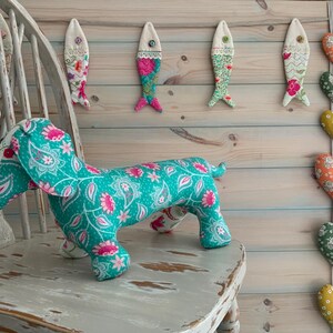 May include: A turquoise dachshund soft toy with a floral pattern sits on a distressed white chair. Fabric fish and heart-shaped ornaments hang on a wooden wall, creating a whimsical, handcrafted theme.