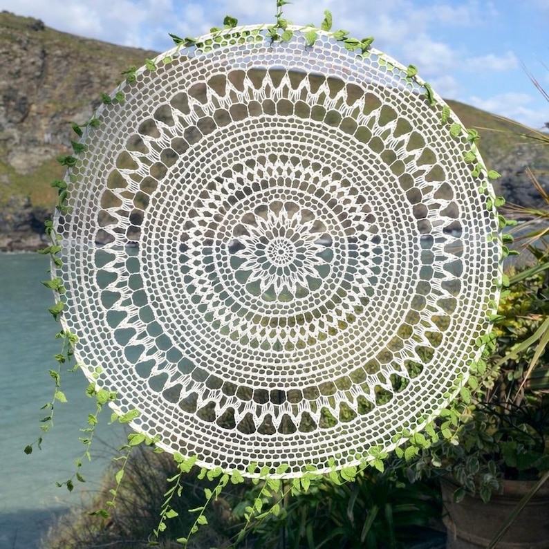 May include: A large, circular white crochet doily with intricate lace patterns. The doily is adorned with small green leaves around the edge. The background shows a coastal landscape with water and hills.