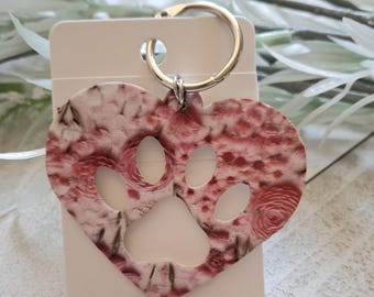 May include: Heart-shaped keychain with a paw print cutout. The keychain features a red and white floral pattern and a silver key ring. The heart is attached to a white card.