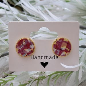 May include: A pair of handmade earrings with a red, white, and yellow patterned design. The earrings are round and have a gold metal setting. The earrings are displayed on a white card with the word "Handmade" written on it.