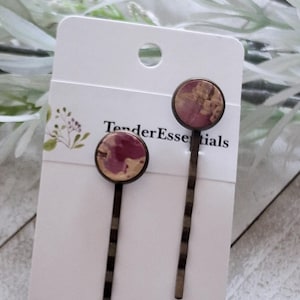 Op de afbeelding: Twee haarspelden met ronde, roze en bruinachtige bloemmotieven. De spelden hebben een donkere metalen afwerking en worden gepresenteerd op een witte kaart met de tekst "TenderEssentials". De achtergrond is vervaagd met groen.