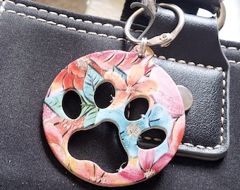 May include: A round, floral-patterned keychain with a paw print cutout. The keychain features a colorful design with red, pink, blue, and green flowers. It is attached to a silver-colored clasp and is hanging on a black bag.