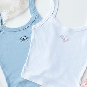 May include: Two tank tops, one light blue and one white, with embroidered initials. The light blue top has black initials, and the white top has pink initials. A small bouquet of pink and white flowers with a white ribbon is in the bottom right corner.