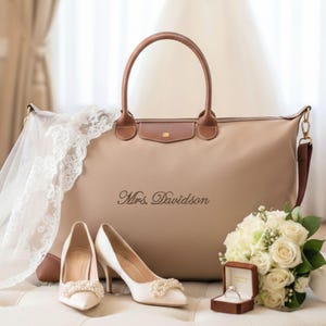 May include: A beige tote bag with brown leather accents, the name "Mrs. Davidson" is printed on the front. A pair of white heels with pearl embellishments, a bouquet of white roses, and a ring box are also present.