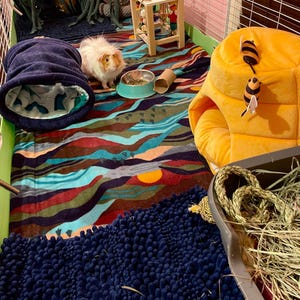 May include: A guinea pig enclosure featuring a colorful landscape-patterned fleece liner, a dark blue fabric tunnel, a yellow beehive-shaped plush hideout with two bee toys, a small wooden stool, a metal food bowl, and a dark blue textured mat.