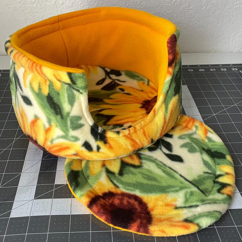 May include: A round, soft pet bed with a sunflower print on a cream background, featuring a bright yellow interior lining and a matching removable circular cushion base. The fabric has a fleece-like texture with green leaves and brown centers.
