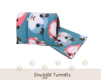 May include: A soft, teal-colored fabric tunnel for small pets, featuring a repeating pattern of white and pink guinea pig faces. The tunnel includes a matching rectangular pee pad. Text on the image reads: Snuggle tunnels, Pee Pad Included | 12 Patterns.