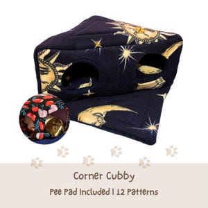 May include: A navy blue corner cubby for small pets, featuring a celestial sun and moon design. The cubby has two circular openings and includes a pee pad. The image also shows a small pet inside a cherry-patterned bed.