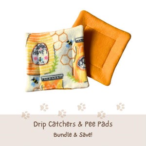 May include: Two square fleece pads. One pad features a cream background with a repeating pattern of honey jars, bees, and honeycomb outlines. The other pad is solid orange. Both pads have stitched edges and a soft, textured fabric surface.