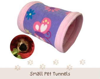 May include: A cylindrical small pet tunnel featuring a purple fleece exterior with a pink, white, and yellow butterfly and flower pattern, finished with solid pink fleece ends. A circular inset shows a guinea pig inside a red fabric tunnel.