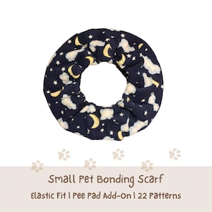 May include: A circular fabric pet bonding scarf featuring a dark navy blue background with a repeating pattern of yellow crescent moons, white clouds, and small white stars. The text below reads: Small Pet Bonding Scarf, Elastic Fit | Pee Pad Add-On | 22 Patterns.