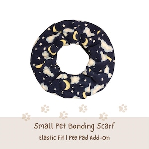 May include: A circular fabric bonding scarf for small pets featuring a dark blue background with a pattern of yellow crescent moons, white stars, and white clouds. The text below reads Small Pet Bonding Scarf, Elastic Fit | Pee Pad Add-On.