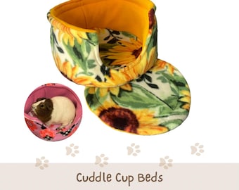 May include: A soft, round pet bed featuring a sunflower print on a white background with a solid yellow interior lining. A separate circular matching pad is included. A small inset shows a brown and white guinea pig resting in a pink floral bed.
