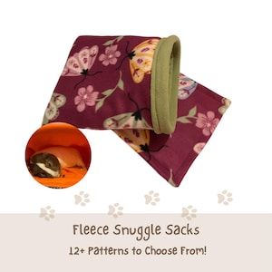 May include: A fleece snuggle sack featuring a dark maroon background with a pattern of light-colored moths and pink flowers, finished with a sage green trim. A circular inset shows a small guinea pig resting inside an orange fleece sack.