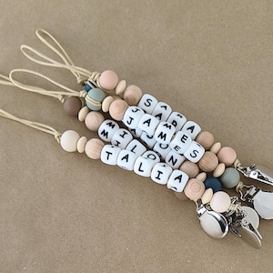 May include: Three dummy clips featuring beige nylon cords, silver metal clasps, and a mix of natural wood and matte silicone beads in shades of tan, blue, and green. Each clip includes white square beads with black letters spelling SARA, JAMES, MILO, and TALIA.