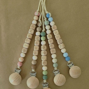 May include: Five dummy clips featuring strings of round silicone and wooden beads in pastel shades of pink, blue, green, and beige. Each clip includes wooden letter beads spelling the names THALIA, MAYA, KADE, MILA, and JASON, finished with a round wooden clip.