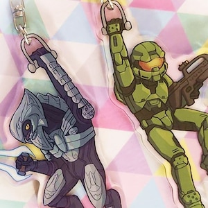 Könnte beinhalten: Zwei Schlüsselanhänger aus Acryl mit Charakteren aus der Videospielserie Halo. Einer zeigt einen grün gepanzerten Spartan mit einer Waffe, der andere einen Covenant Elite mit einem blauen Energieschwert. Beide Schlüsselanhänger haben silberfarbene Hardware.