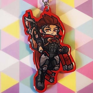 Puede incluir: Un llavero con un personaje chibi estilizado de cabello castaño, que viste un traje táctico gris oscuro y negro con una bufanda roja, sosteniendo un arma futurista grande. El personaje tiene un borde rojo y está unido a una anilla de metal plateado.