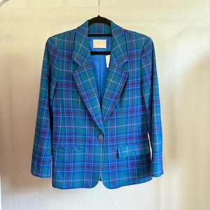 K&ouml;nnte beinhalten: Ein Vintage-Blazer mit Karomuster in T&uuml;rkis-, Blau- und Lilat&ouml;nen, der an einer wei&szlig;en Stange h&auml;ngt. Der Blazer hat einen Ein-Knopf-Verschluss, gekerbte Revers und zwei Vordertaschen. Das Innenfutter ist einfarbig blau.