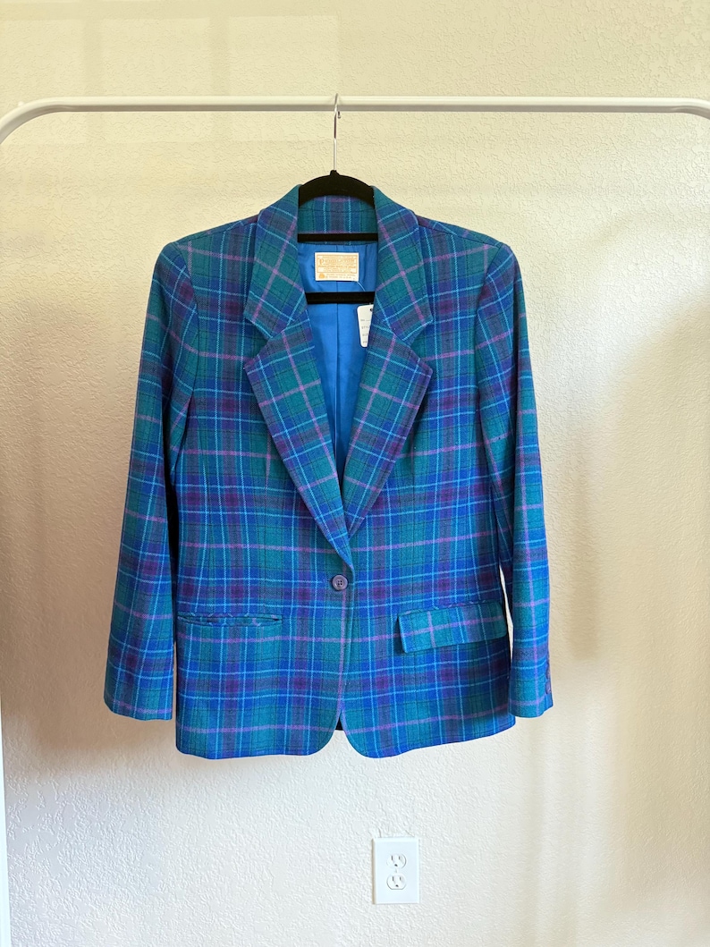 K&ouml;nnte beinhalten: Ein Vintage-Blazer mit Karomuster in T&uuml;rkis-, Blau- und Lilat&ouml;nen, der an einer wei&szlig;en Stange h&auml;ngt. Der Blazer hat einen Ein-Knopf-Verschluss, gekerbte Revers und zwei Vordertaschen. Das Innenfutter ist einfarbig blau.