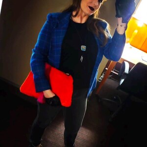 K&ouml;nnte beinhalten: Eine Person tr&auml;gt einen blau karierten Blazer &uuml;ber einem schwarzen Oberteil und einer schwarzen Hose und h&auml;lt eine leuchtend rote Clutch. Das Outfit ist mit einer silbernen Halskette und schwarzen Stiefeln mit gelben Schn&uuml;rsenkeln ausgestattet. Der Hintergrund ist ein dunkles Interieur.