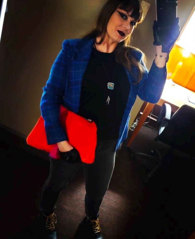 K&ouml;nnte beinhalten: Eine Person tr&auml;gt einen blau karierten Blazer &uuml;ber einem schwarzen Oberteil und einer schwarzen Hose und h&auml;lt eine leuchtend rote Clutch. Das Outfit ist mit einer silbernen Halskette und schwarzen Stiefeln mit gelben Schn&uuml;rsenkeln ausgestattet. Der Hintergrund ist ein dunkles Interieur.