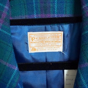 K&ouml;nnte beinhalten: Nahaufnahme eines Vintage-Pendleton-Wollblazers in einem teal-, lila- und blau karierten Muster. Das Innenfutter ist leuchtend blau, mit einem Pendleton-Etikett sichtbar. Der Blazer h&auml;ngt an einem schwarzen Kleiderb&uuml;gel.