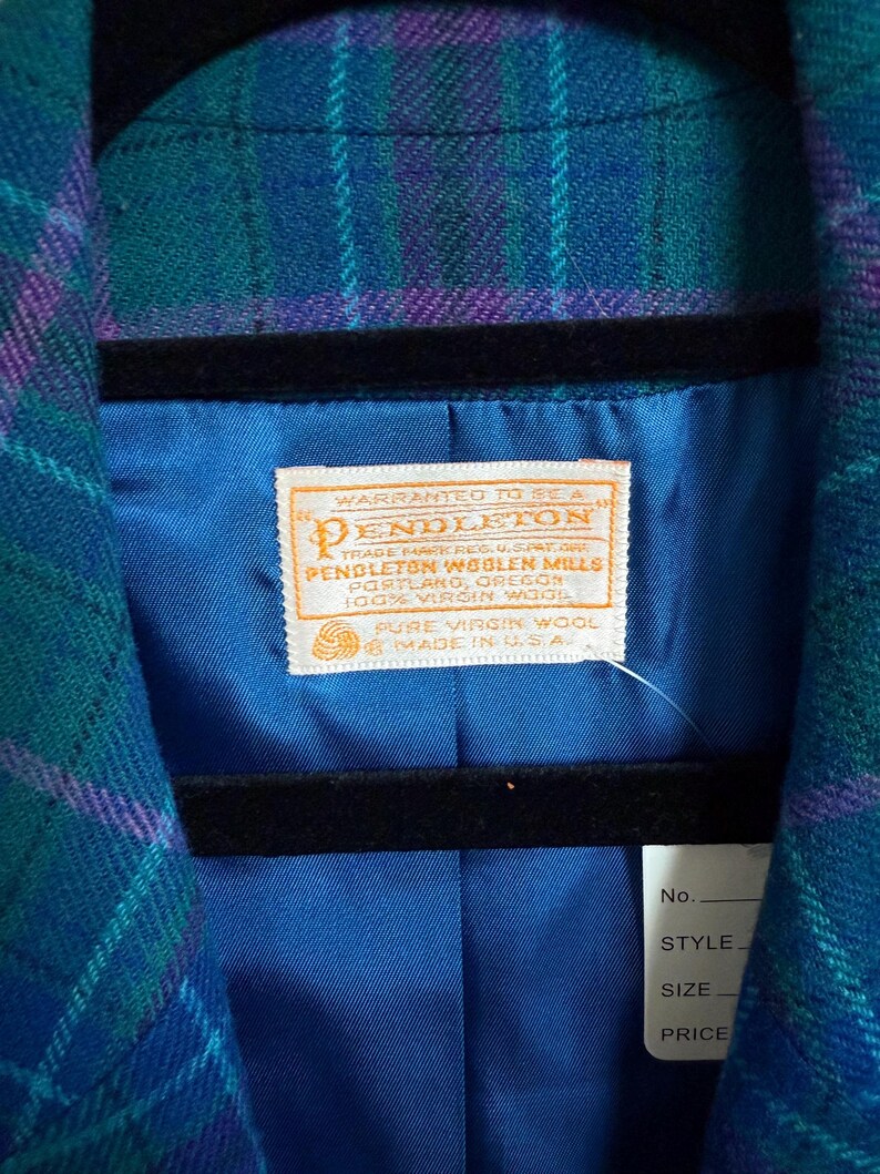 K&ouml;nnte beinhalten: Nahaufnahme eines Vintage-Pendleton-Wollblazers in einem teal-, lila- und blau karierten Muster. Das Innenfutter ist leuchtend blau, mit einem Pendleton-Etikett sichtbar. Der Blazer h&auml;ngt an einem schwarzen Kleiderb&uuml;gel.