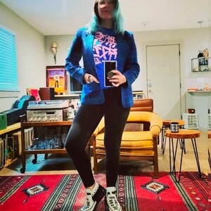 K&ouml;nnte beinhalten: Blauer Blazer mit Karomuster &uuml;ber einem blauen T-Shirt mit rosa japanischen Schriftzeichen. Die Person tr&auml;gt schwarze Leggings und wei&szlig;-schwarze Schuhe im Mokassin-Stil. Der Teppich ist rot mit einem geometrischen Muster.