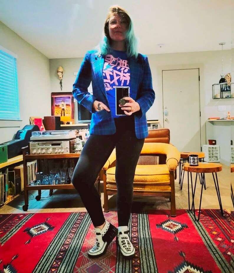 K&ouml;nnte beinhalten: Blauer Blazer mit Karomuster &uuml;ber einem blauen T-Shirt mit rosa japanischen Schriftzeichen. Die Person tr&auml;gt schwarze Leggings und wei&szlig;-schwarze Schuhe im Mokassin-Stil. Der Teppich ist rot mit einem geometrischen Muster.