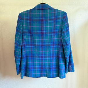 K&ouml;nnte beinhalten: Blazer mit blauem, t&uuml;rkisem und lila Karomuster, der an einem schwarzen Kleiderb&uuml;gel h&auml;ngt. Der Blazer hat ein klassisches Design mit einem Reverskragen und gekn&ouml;pften Manschetten. Das Karomuster ist eine Mischung aus T&uuml;rkis-, Blau- und Lilat&ouml;nen.
