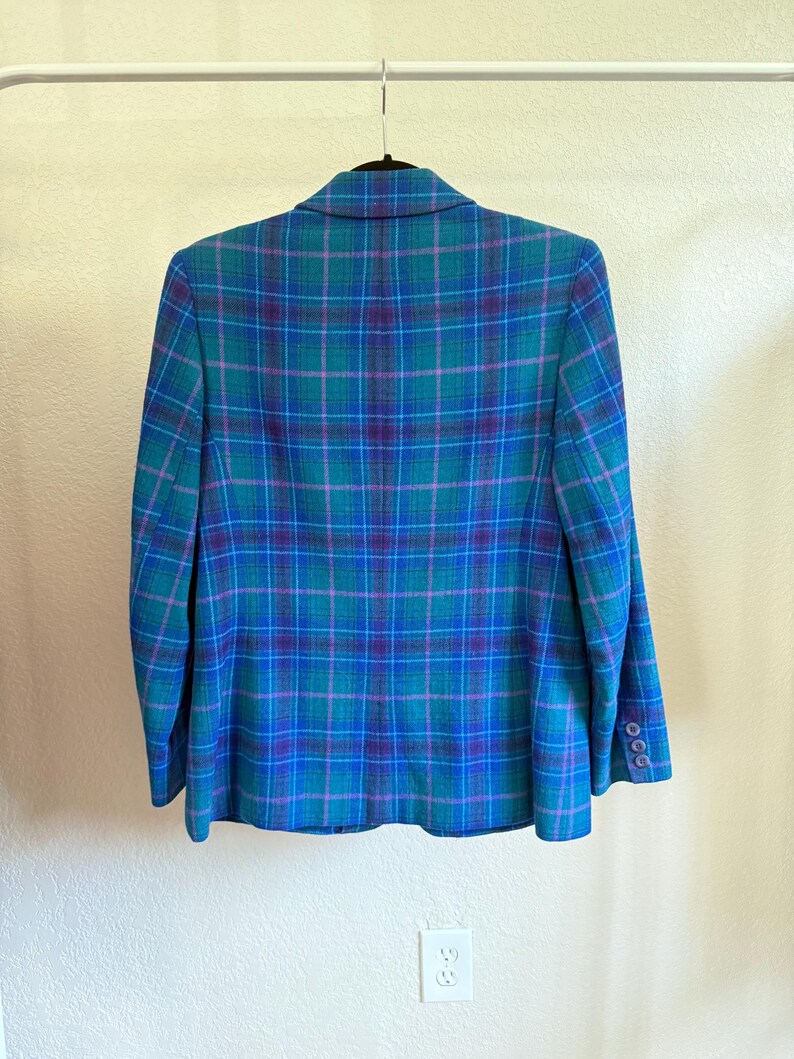 K&ouml;nnte beinhalten: Blazer mit blauem, t&uuml;rkisem und lila Karomuster, der an einem schwarzen Kleiderb&uuml;gel h&auml;ngt. Der Blazer hat ein klassisches Design mit einem Reverskragen und gekn&ouml;pften Manschetten. Das Karomuster ist eine Mischung aus T&uuml;rkis-, Blau- und Lilat&ouml;nen.