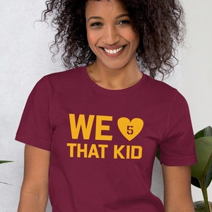 May include: Burgundy t-shirt with the text "WE ♥ THAT KID" in gold. The heart symbol contains the number 5. The shirt is a short-sleeved crew neck style.