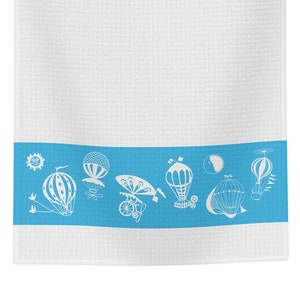 May include: White kitchen towel with a blue border featuring a pattern of hot air balloons. The balloons are white and have a whimsical design.