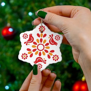 May include: A white ceramic snowflake ornament with a red and yellow geometric design featuring stylized birds. The ornament is being held by a hand.