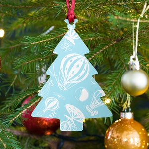 May include: A blue Christmas tree ornament with a white design of hot air balloons and other whimsical elements. The ornament is hanging on a green Christmas tree with red and gold ornaments.
