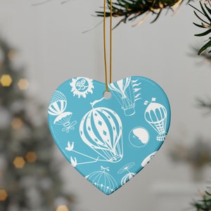 May include: A heart-shaped Christmas ornament with a light blue background and a white hot air balloon pattern. The ornament is hanging from a gold string and is displayed on a Christmas tree. The ornament has a glossy finish.