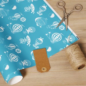 May include: A roll of blue wrapping paper with a white hot air balloon pattern, a brown tag, scissors, and twine on a wooden surface. The paper features various hot air balloon designs and other whimsical elements.